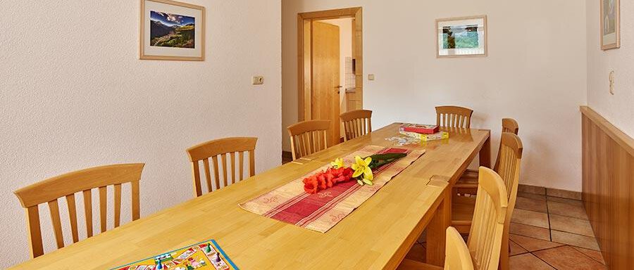 A bright dining area with a long wooden table and several chairs. On the table are flowers and a board game. - Pension Gritsch Ewald - Sölden