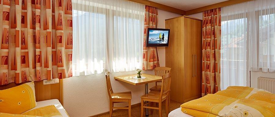 A cozy room with two beds and colorful curtains. There is a table, a television, and plenty of daylight. - Pension Gritsch Ewald - Sölden