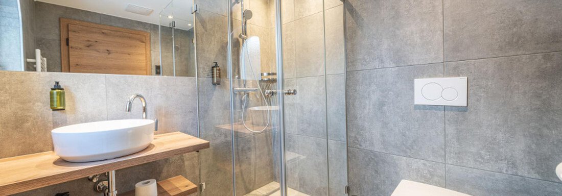 Badezimmer DZ Arnika | © Pension Miramonte (CC C) A modern bathroom with a glass shower cabin and an elegant bathtub. The walls are designed with gray tiles, and there are wooden details. - Pension Miramonte - Sölden