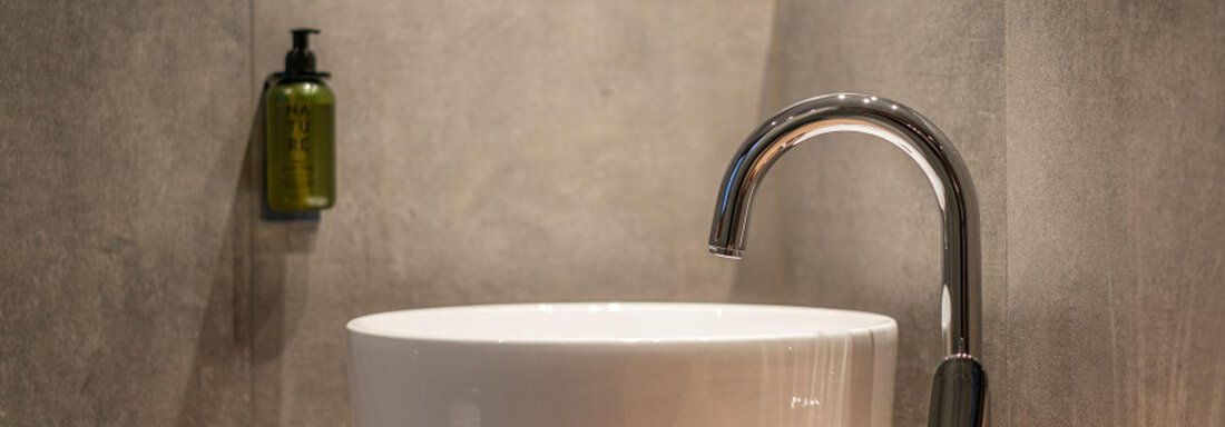 A modern ceramic sink sits on a wooden shelf. In the background, gray tiles and a soap dispenser are visible. - Pension Miramonte - Sölden