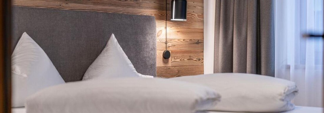 A cozy bedroom with a double bed and decorative pillows. The wall is made of wood, and there is a modern lamp next to the bed. - Pension Miramonte - Sölden