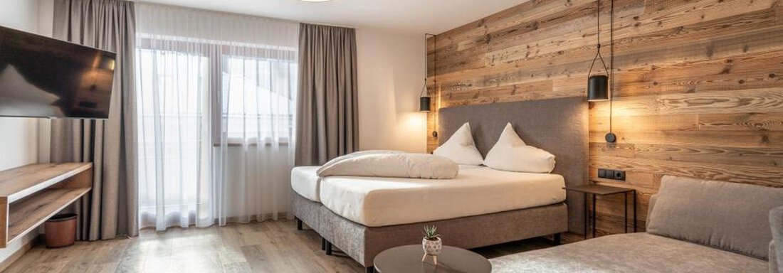 A modern hotel room with a large bed and a comfortable armchair. The wood paneling on the wall and the soft lighting create a cozy atmosphere. - Pension Miramonte - Sölden