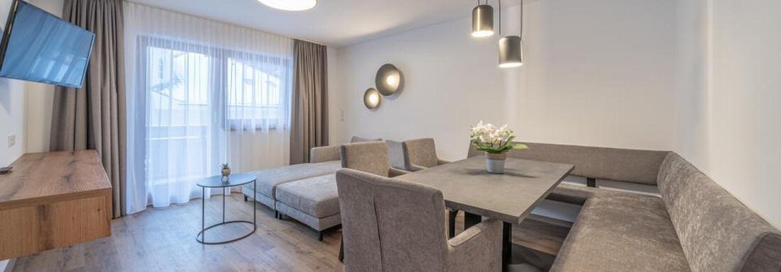 A modern living room with a cozy dining area and a large window. The decor is bright and inviting, with a television on the wall. - Pension Miramonte - Sölden