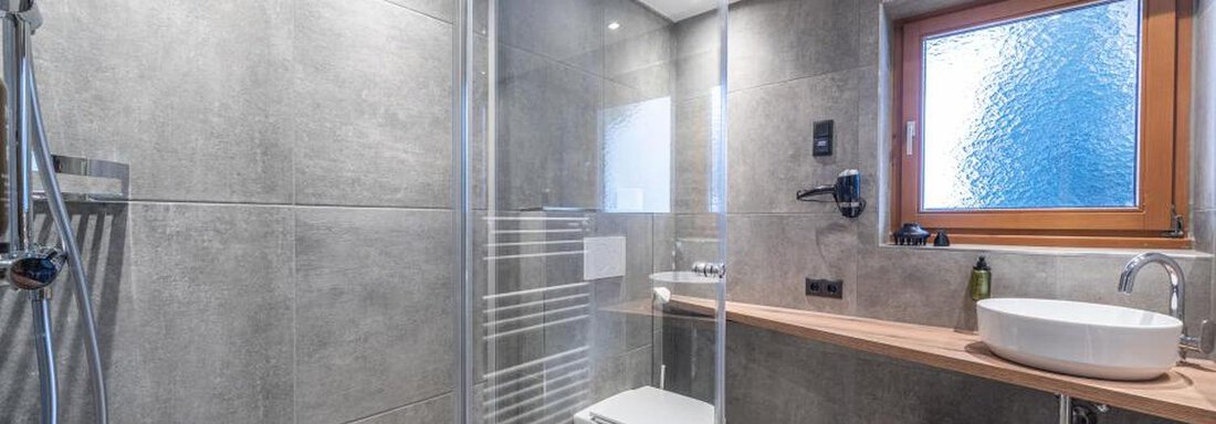 A modern bathroom with gray tiles and a glass shower. It is bright and stylishly furnished with a wooden vanity and a large window. - Pension Miramonte - Sölden