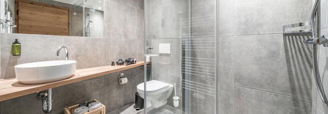 A modern bathroom with gray tiles and a walk-in shower. On the sink, there is a stylish bowl and a wooden shelf. - Pension Miramonte - Sölden