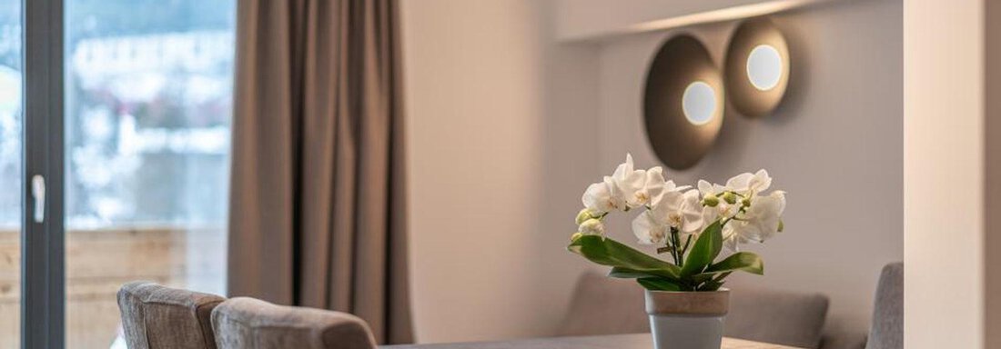 A modern living room with a wooden table and three chairs. On the table is a pot with white flowers, and large windows let in plenty of light. - Pension Miramonte - Sölden