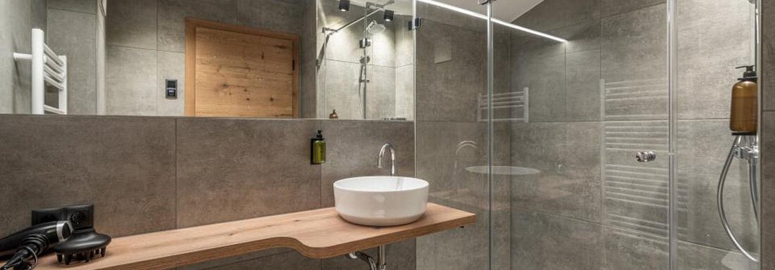 A modern bathroom with an elegant glass shower and gray tiles. A simple wooden sink and a large mirror complete the design. - Pension Miramonte - Sölden