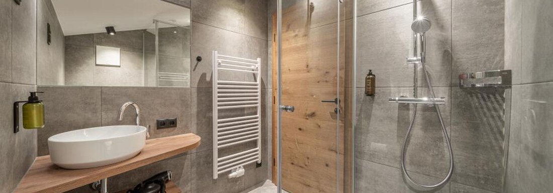 A modern bathroom with a glass shower and a sink with a wooden shelf. The walls are in neutral gray tones. - Pension Miramonte - Sölden