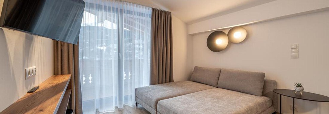 A modern living and bedroom with a comfortable sofa and a television on the wall. Large windows with curtains let in plenty of daylight. - Pension Miramonte - Sölden