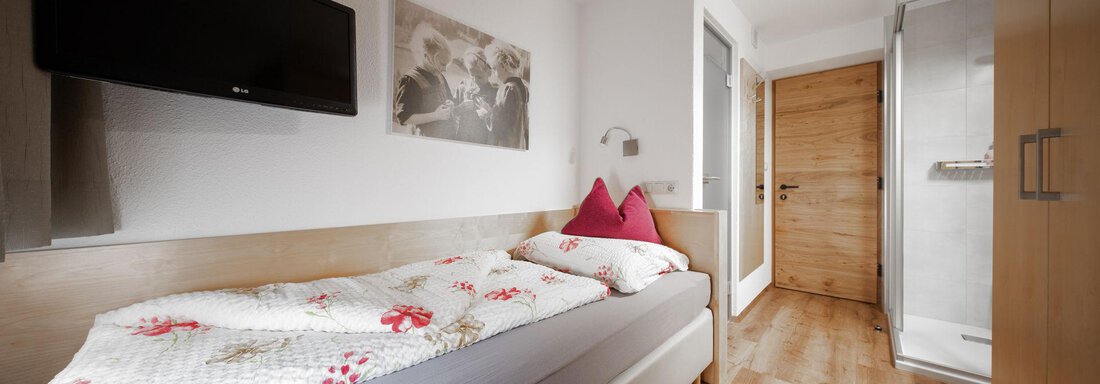 A cozy bedroom with a single bed and a television on the wall. The room has a wooden floor and a window that leads to a bathroom. - Pension Pitze - Sölden