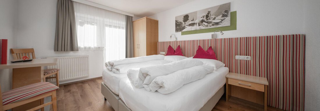 A modern bedroom with two single beds and colorful pillows. The room has hardwood floors and plenty of natural light through a large window. - Pension Pitze - Sölden