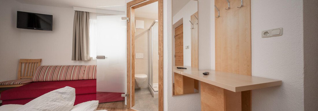 A modern room with wooden flooring and a large window. There is a bed, a couch, as well as a table with a mirror and coat hooks. - Pension Pitze - Sölden