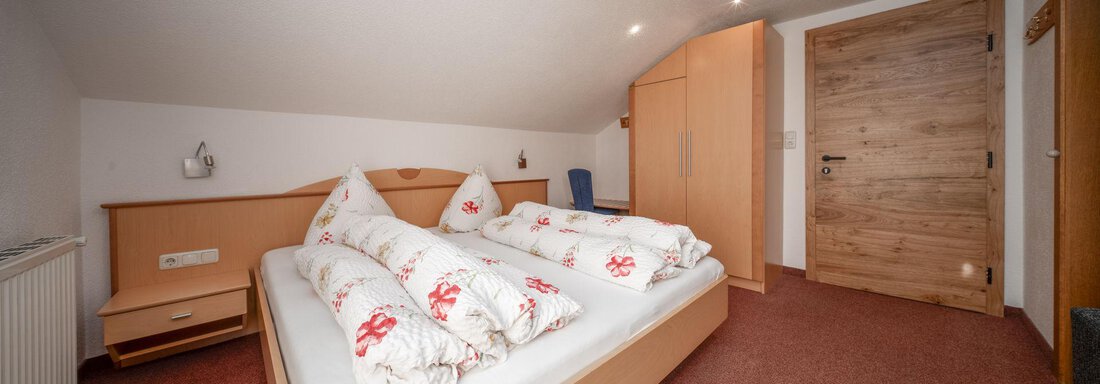 A cozy bedroom with a large bed and red floral patterns. The room has light walls and wooden details. - Pension Pitze - Sölden