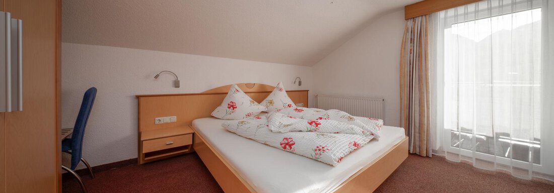 A bright bedroom with a large bed and white bedcovers. The sun is shining through the windows, creating a pleasant atmosphere. - Pension Pitze - Sölden