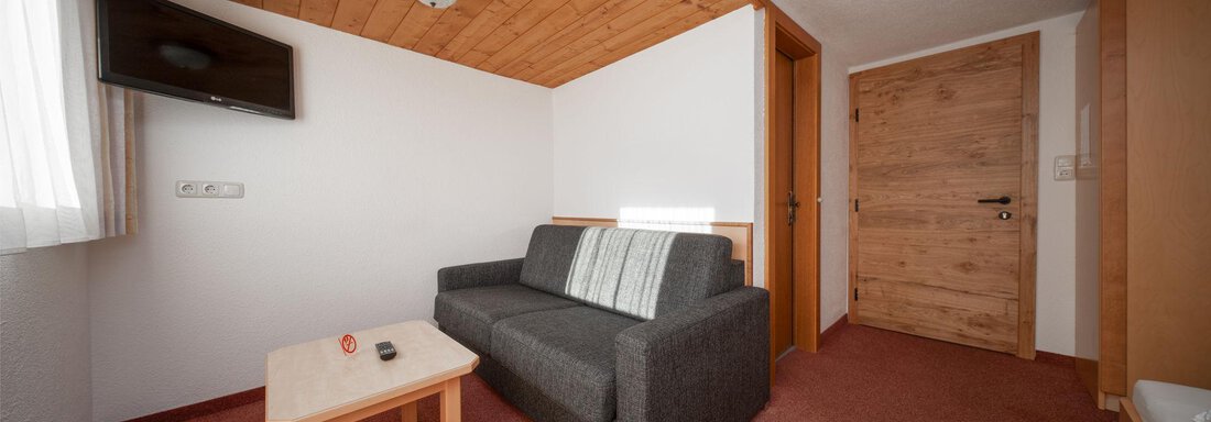 A cozy room with a gray sofa and a small table. On the wall, there is a television and a window lets in a lot of light. - Pension Pitze - Sölden