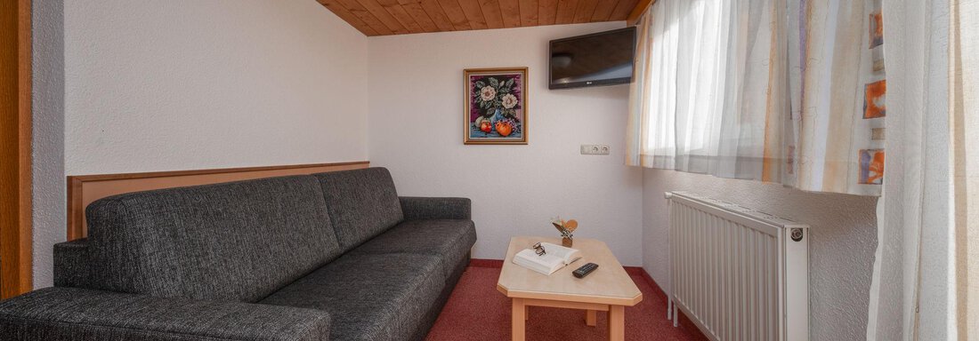 A cozy room with a gray sofa and a small table. On the wall, there is a television and a window with curtains. - Pension Pitze - Sölden