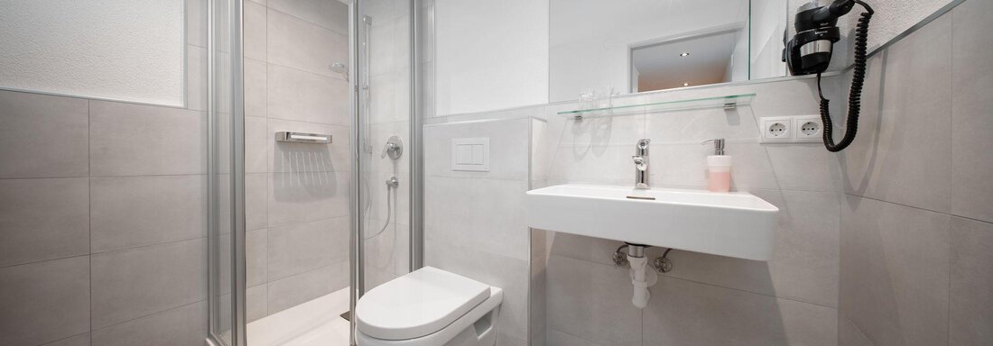 A modern bathroom with a shower, a sink, and a mirror. The walls are tiled in light gray, and there is a telephone system on the wall. - Pension Pitze - Sölden