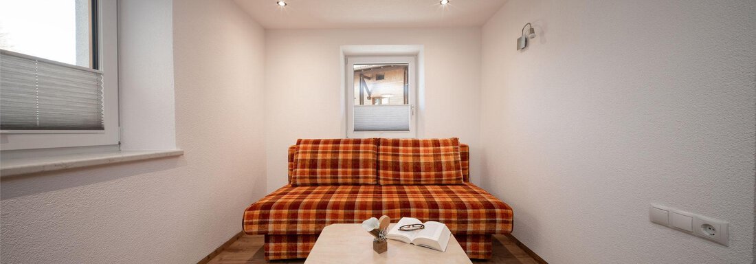 A cozy room with a checkered sofa and a small table. There is a window that provides natural light. - Pension Pitze - Sölden