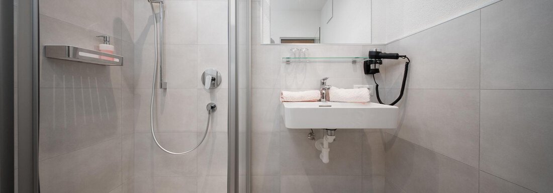 A modern bathroom with a shower and sink. The walls are light and simply done in gray. - Pension Pitze - Sölden