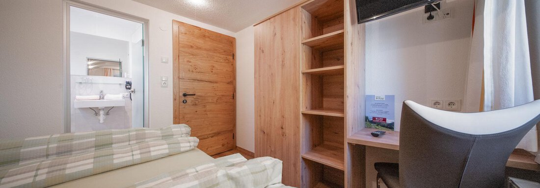 A cozy room with wooden interior and a television on the wall. There is a bed and a desk as well as access to a bathroom. - Pension Pitze - Sölden