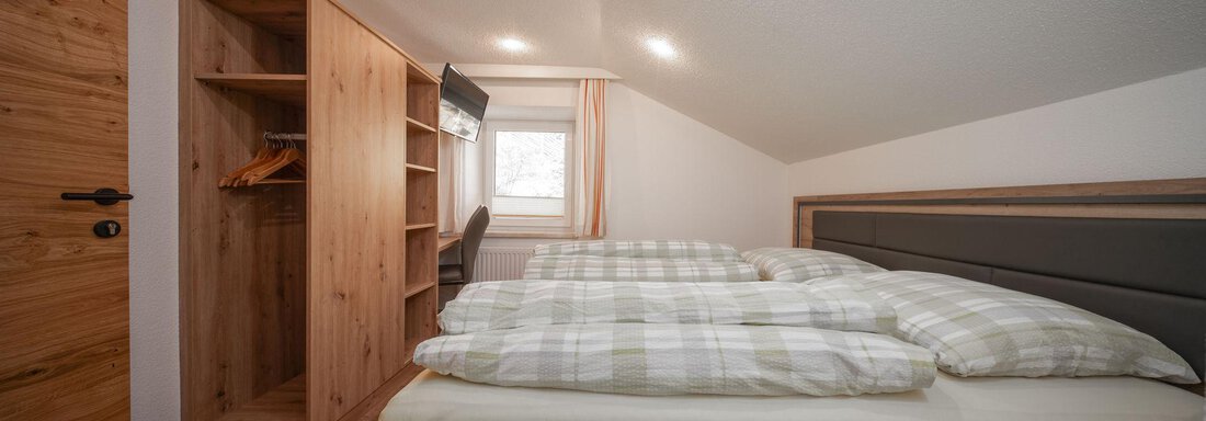 A cozy bedroom with two beds and a wooden wardrobe. The room has light walls and a large window that lets in daylight. - Pension Pitze - Sölden