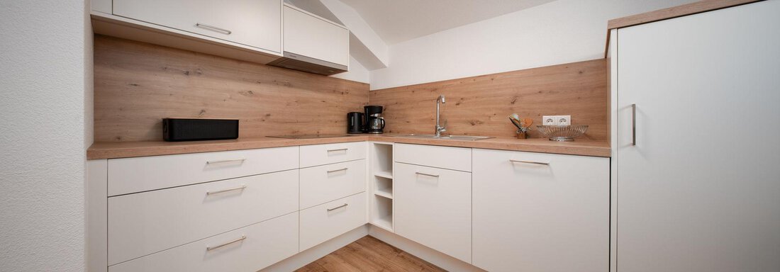 A modern kitchen with white furniture and wooden shelves. The floor is made of wood and there is a sink as well as kitchen appliances. - Pension Pitze - Sölden