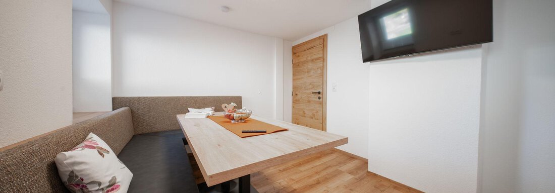 A modern dining area with a wooden table and comfortable benches. The walls are light and a television is mounted on the wall. - Pension Pitze - Sölden