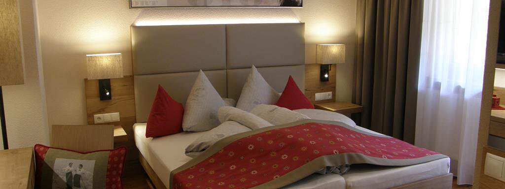 A cozy hotel room with a large bed, red pillows, and modern wooden furnishings. Large windows let in plenty of light and create an inviting atmosphere. - Pension Sportalm - Sölden