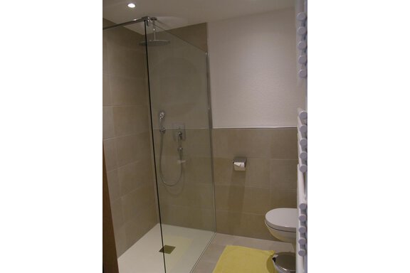 A modern bathroom with a glass shower and a wall-mounted toilet. The walls are in neutral shades, and a yellow towel is lying on the floor. - Pension Sportalm - Sölden