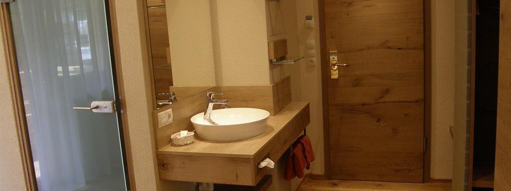A modern bathroom with wooden details and a generous mirror surface. The door is made of wood and the floor is laid with warm wood. - Pension Sportalm - Sölden