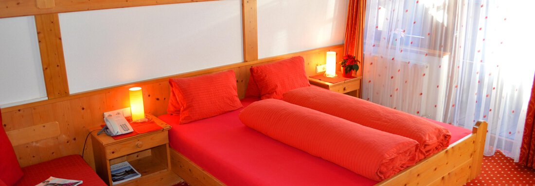 Double room with balcony A cozy room with two red beds and a wooden frame. The windows are decorated with curtains, and there is a seating area next to the bed. - Pension "Zur alten Mühle" - Sölden