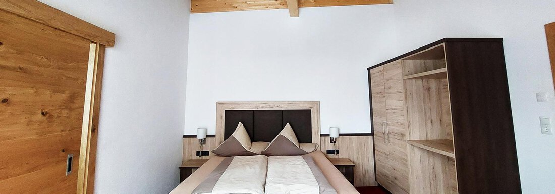 A modern bedroom with a large bed and wooden ceiling. There is a wardrobe and a beautiful red carpet floor. - Pension "Zur alten Mühle" - Sölden