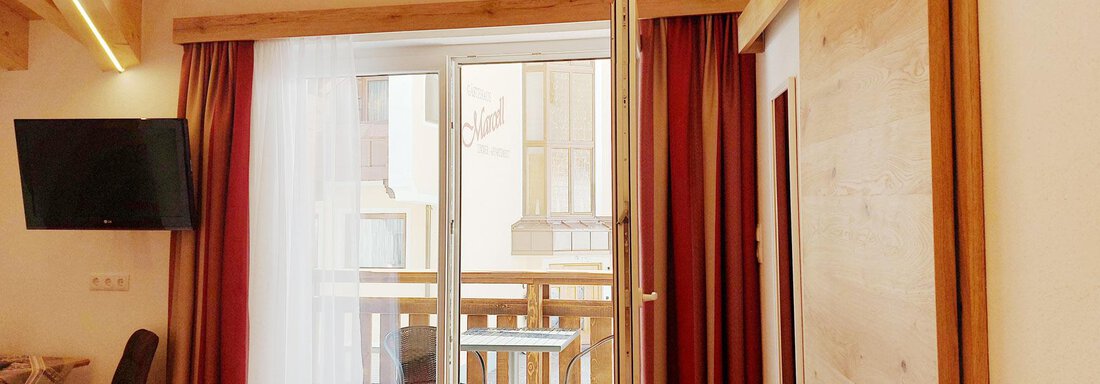 A cozy room with wooden elements and a balcony. A television and a simple chair are also visible. - Pension "Zur alten Mühle" - Sölden