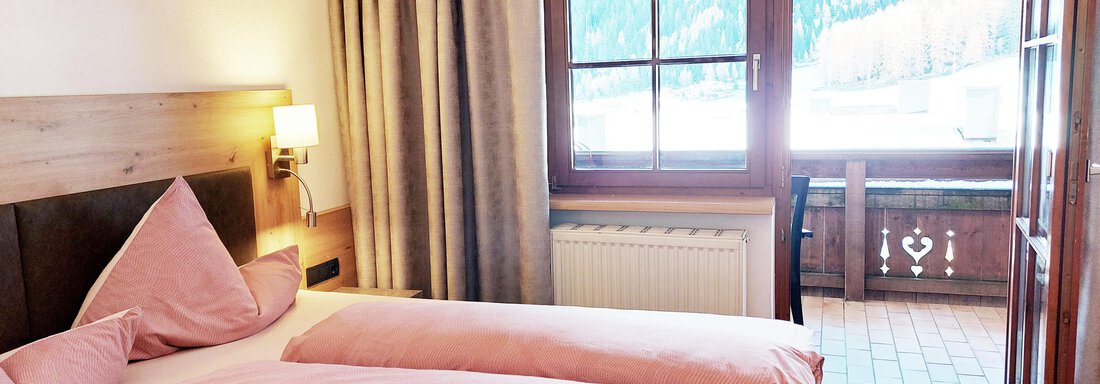 A cozy bedroom with two beds and pink bed linen. The room has large windows overlooking a balcony and the surrounding mountains. - Pension "Zur alten Mühle" - Sölden