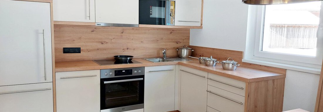 A modern kitchen with wooden paneling and light cabinets. It is well-equipped with a stove, a refrigerator, and a window for plenty of light. - Pension "Zur alten Mühle" - Sölden