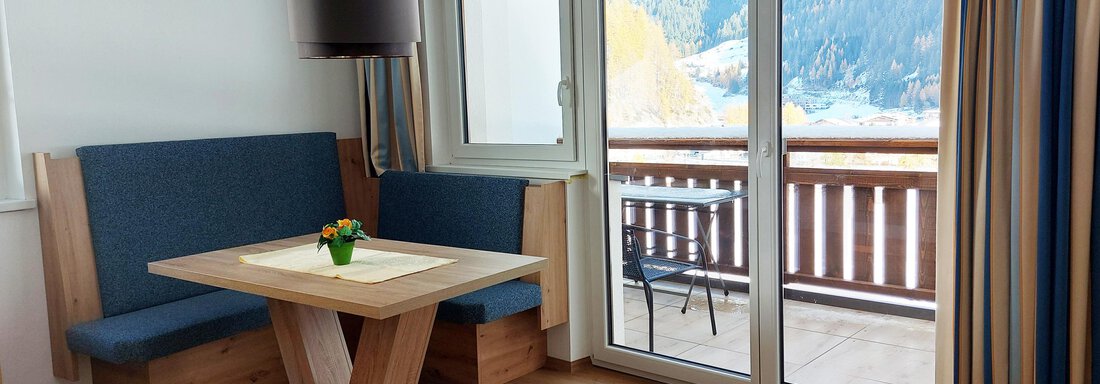 A cozy dining area with a table and comfortable benches. Large windows offer a view of the balcony and the surrounding landscape. - Pension "Zur alten Mühle" - Sölden