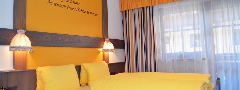A cozy bedroom with a yellow bed and a modern lamp. The walls are decorated in light yellow with a decorative lettering. - Pension "Zur alten Mühle" - Sölden