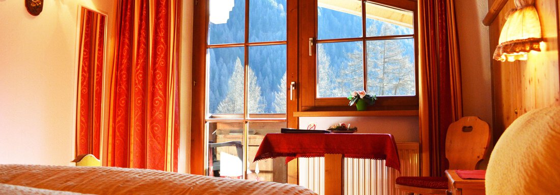 A cozy room with a large window and a view of the mountains. The decor is warm and inviting, with orange curtains and beds. - Pension "Zur alten Mühle" - Sölden