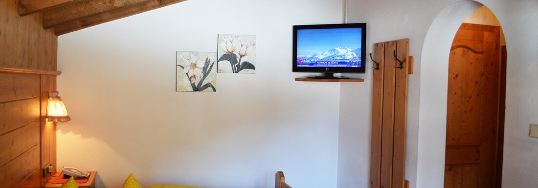 A cozy room with a sloped ceiling, equipped with a television and a large bed. On the bed are yellow pillows and there is a door that leads to another room. - Pension "Zur alten Mühle" - Sölden