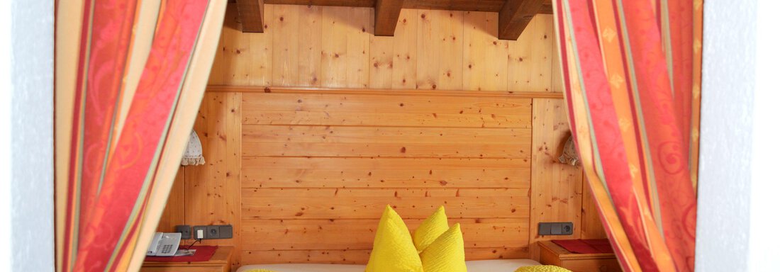 A cozy bedroom with wood paneling and a wooden ceiling beam. The yellow cushions and red curtains add a warm touch to the room. - Pension "Zur alten Mühle" - Sölden