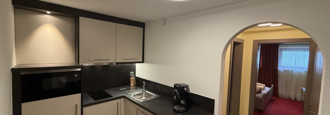 Wohnküche mit Blick ins Schlafzimmer A modern kitchen with black and light cabinets and modern lighting. The living room is visible through an arch. - Pension "Zur alten Mühle" - Sölden