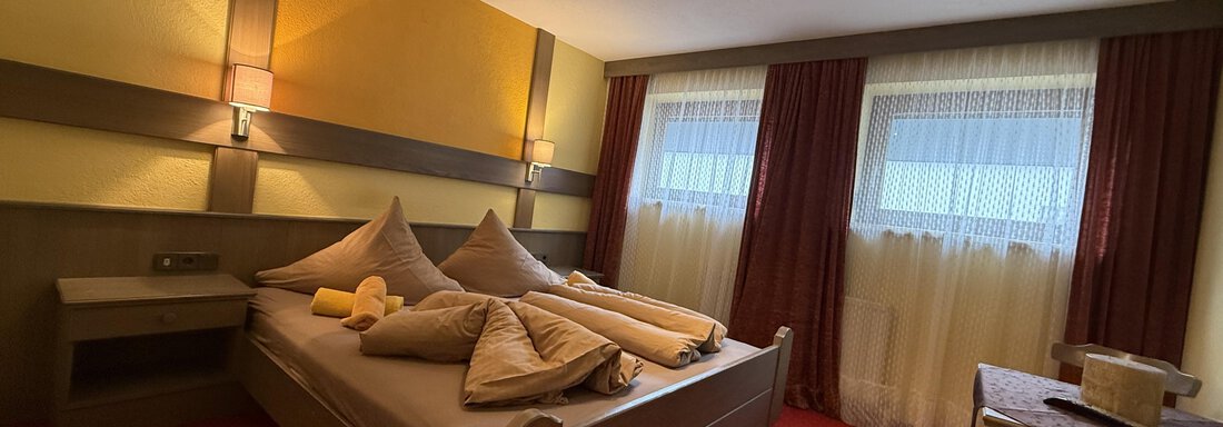 A cozy bedroom with two beds and large pillows. The walls are in shades of yellow and gray, and there is a beautiful view through the windows. - Pension "Zur alten Mühle" - Sölden