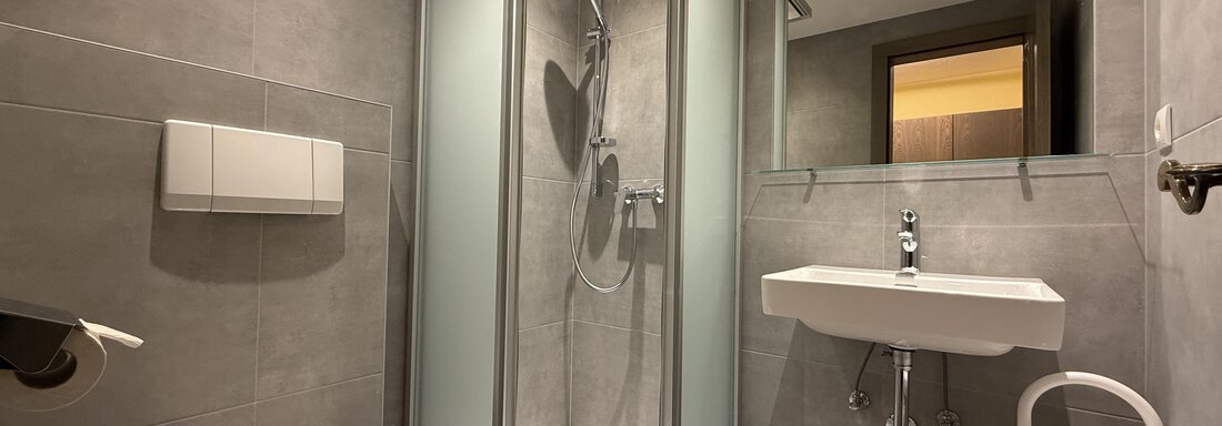 A modern bathroom with gray tiles and a shower. There is a sink and a toilet, all well-lit. - Pension "Zur alten Mühle" - Sölden
