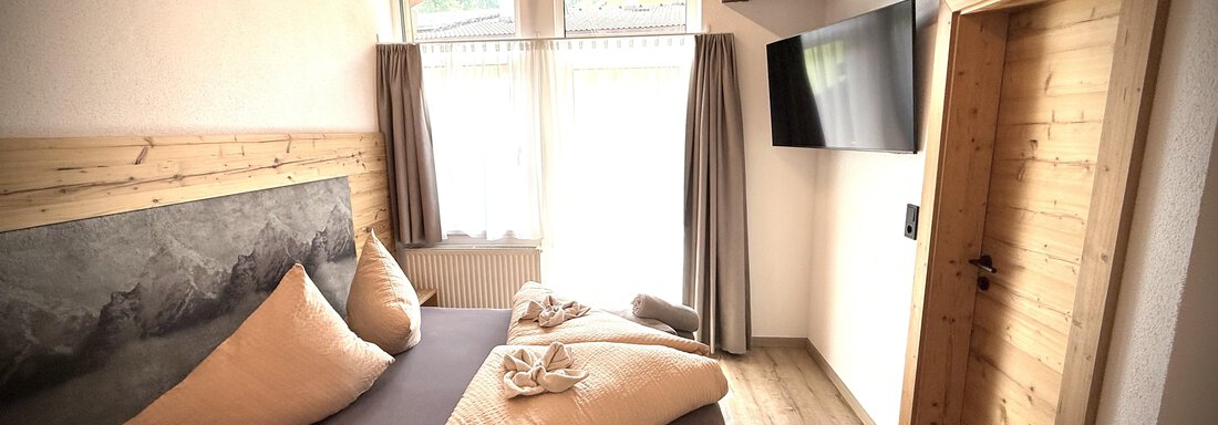 A modern bedroom with wooden decor and large windows. There is a comfortable bed with large pillows and a television on the wall. - Pension "Zur alten Mühle" - Sölden