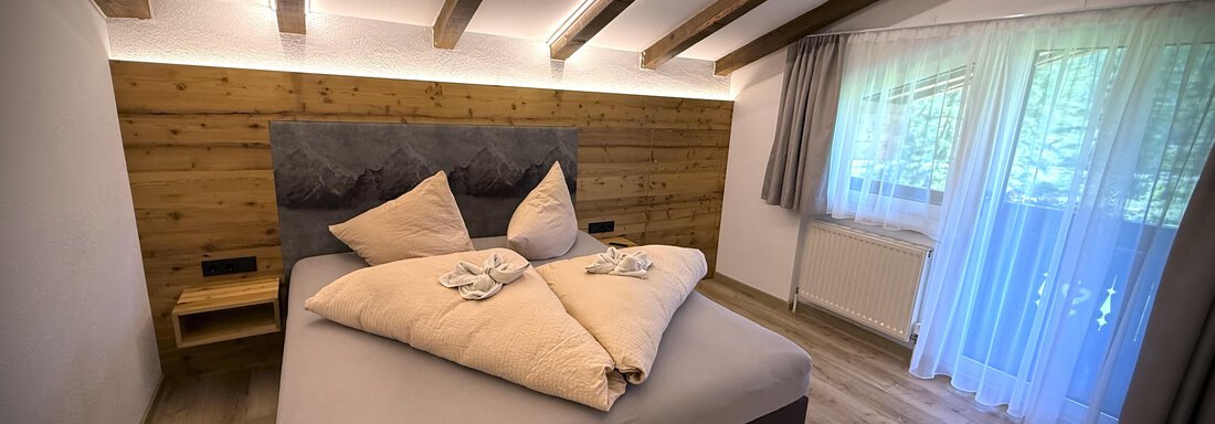 A modern bedroom with a large bed and wood paneling. Bright windows let in plenty of light and create a cozy atmosphere. - Pension "Zur alten Mühle" - Sölden