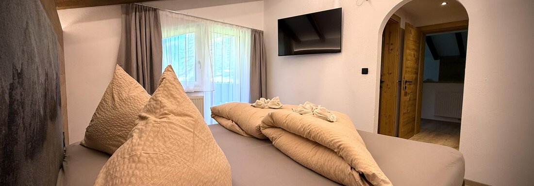 A cozy bedroom with a large bed and many pillows. There is a window with curtains and a television on the wall. - Pension "Zur alten Mühle" - Sölden