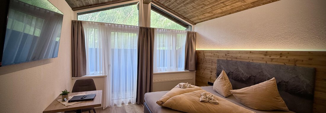 A modern bedroom with a wooden ceiling and large windows. It is bright and cozy with a large bed and a small workspace. - Pension "Zur alten Mühle" - Sölden