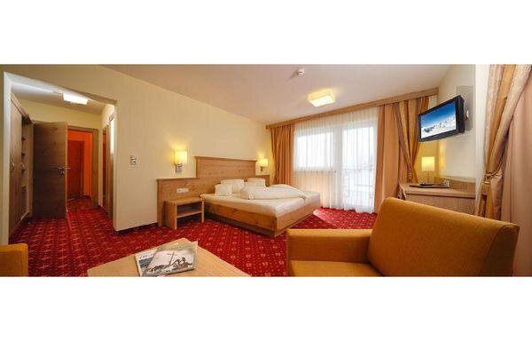A cozy hotel room with a large bed, a small table, and a television. The windows let in a lot of light, and the carpet has a red pattern. - Residenz Allegra - Sölden