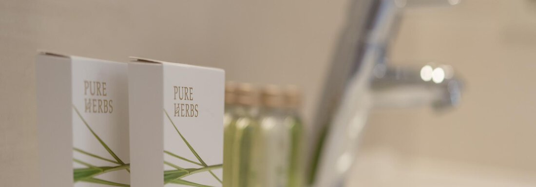 Two soap packages with the inscription "PURE HERBS" are placed next to small bottles on a sink. The background is designed to be light and modern. - Residenz Allegra - Sölden