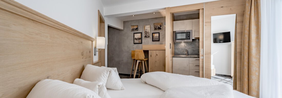 A modern hotel room with a wooden bed and large pillows. In the background, there is a small kitchenette and a television. - Residenz Allegra - Sölden